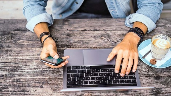Image of computer with one hand on it and one hand holding a cell phone