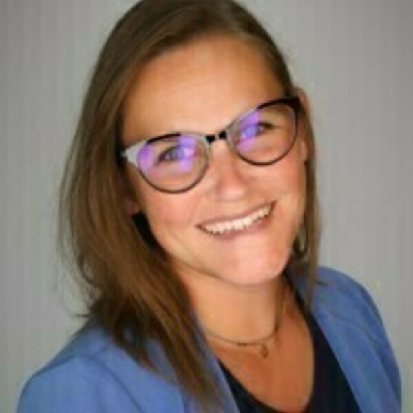 A smiling woman with shoulder-length brown hair and black-rimmed glasses wears a blue blazer and a delicate gold necklace.