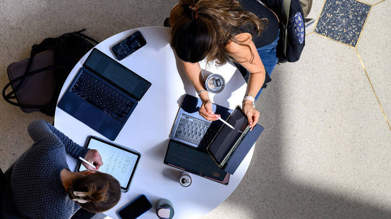 Two students with dark hair work at a white table. One writes math equations on a tablet; the other uses a stylus and laptop.
